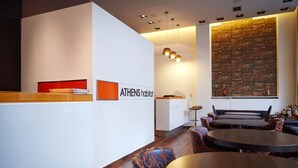 A modern reception area with a white wall and a sign reading "ATHENS habitat". There are wooden tables and chairs, and a large wall with a brick pattern. - Athens Habitat (Marousi)