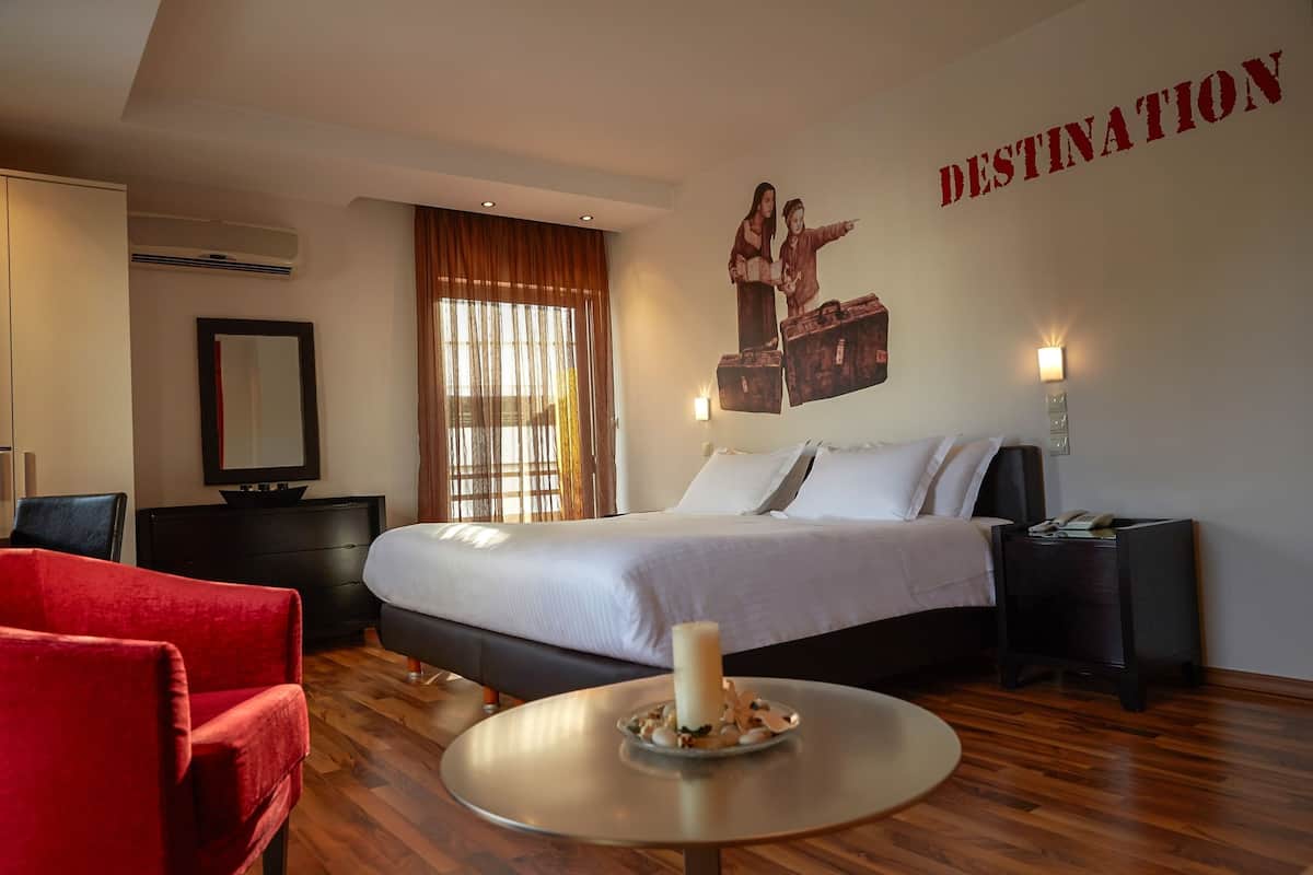 Une chambre d’hôtel avec un lit, une chaise rouge, une table ronde avec une bougie, un téléviseur fixé au mur et un panneau indiquant « DESTINATION ».