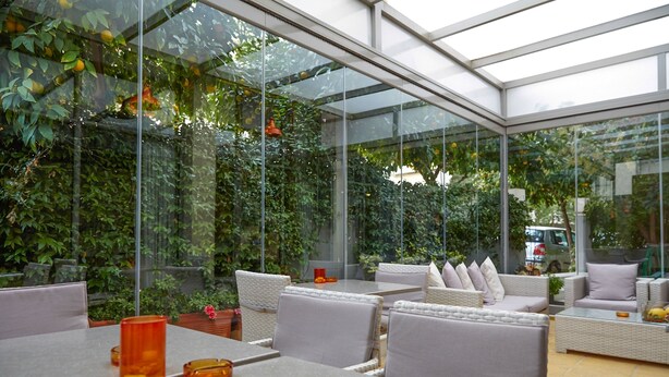 A modern outdoor seating area with glass walls, wicker furniture, and a view of lush greenery. - Athens Habitat (Marousi)
