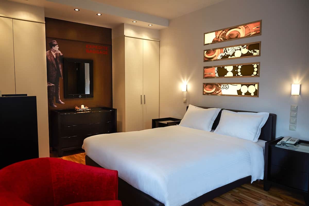 Une chambre d’hôtel avec un grand lit, un fauteuil rouge, une armoire, une commode, un miroir et des œuvres d’art accrochées au mur.