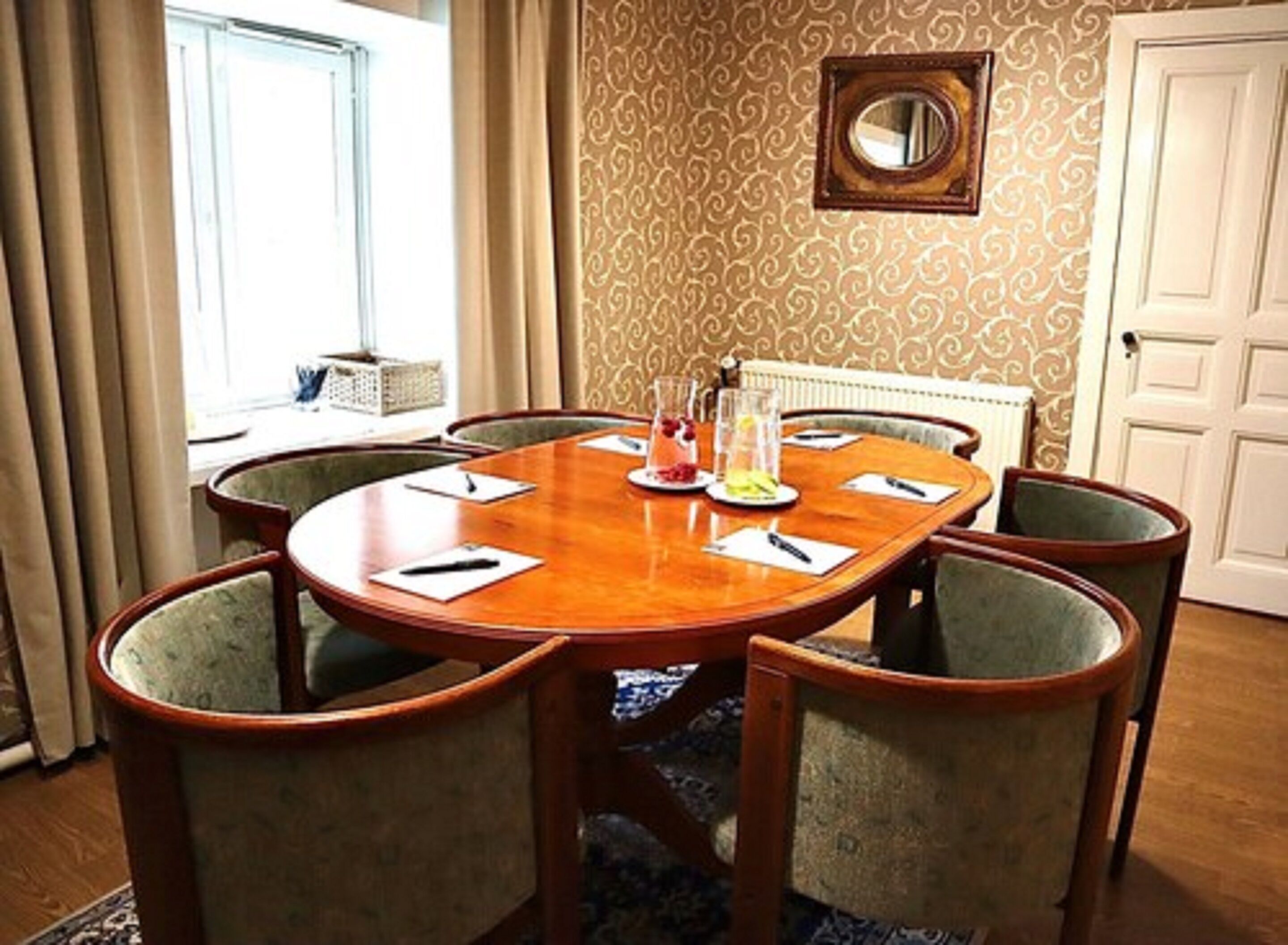 A conference room with a round wooden table, six upholstered chairs, and a decorative mirror on the wall.