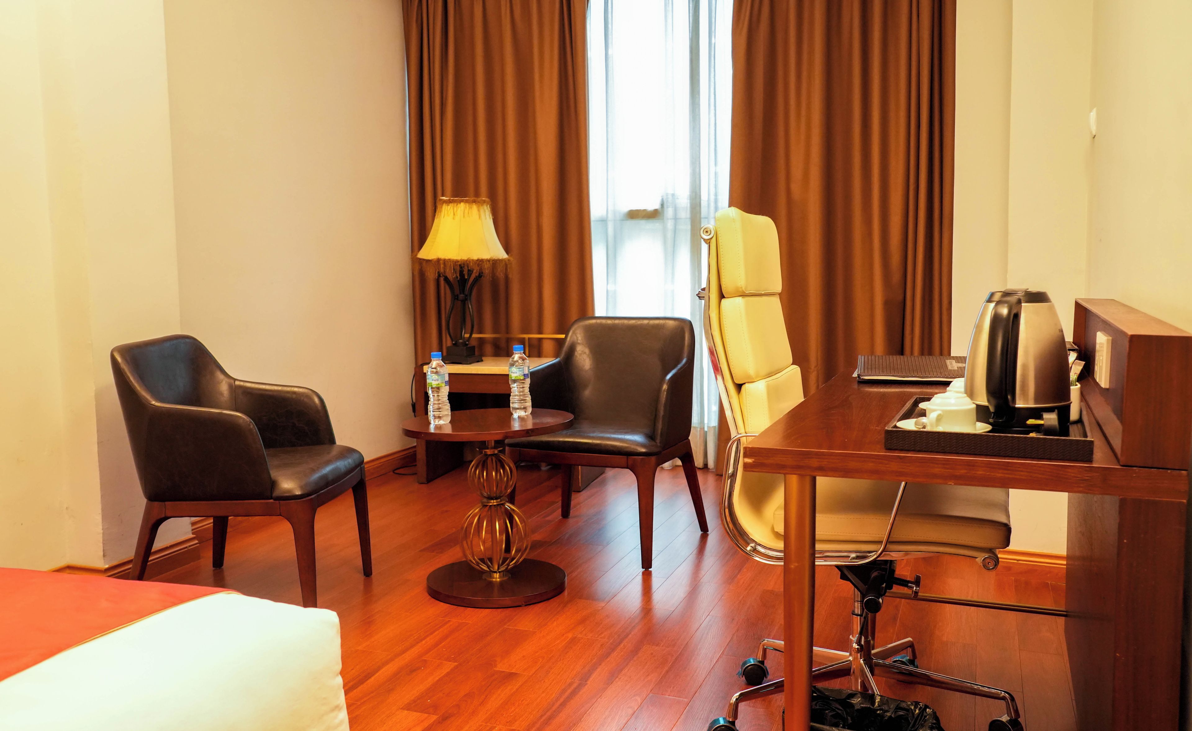 A hotel room with a bed, a desk with a coffee maker, two chairs, a lamp, and a bottle of water.