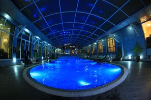 An indoor swimming pool with a glass ceiling, surrounded by lounge chairs and potted plants.