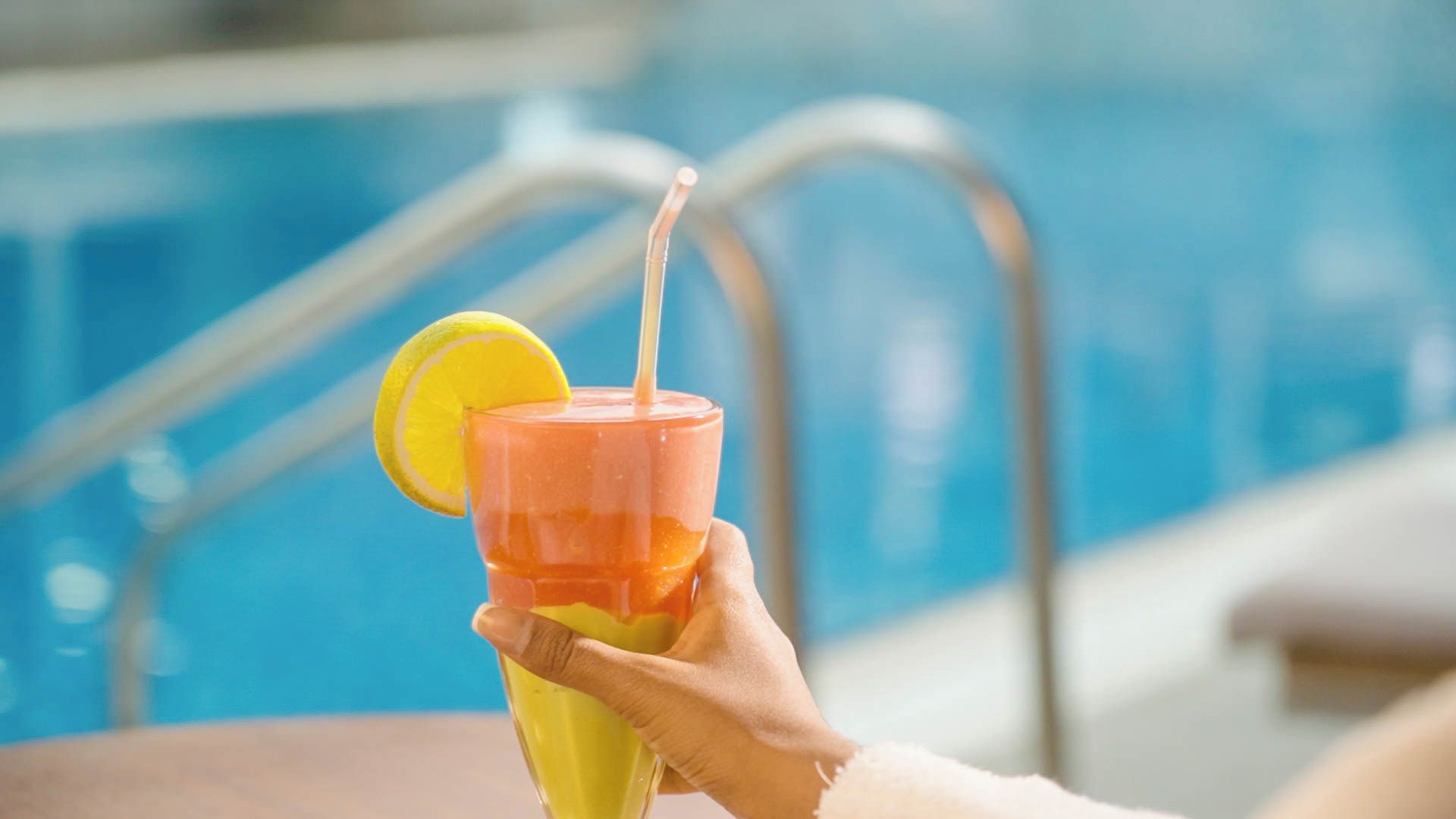 A hand holding a glass with a straw, a slice of lemon, and a drink, by a poolside.