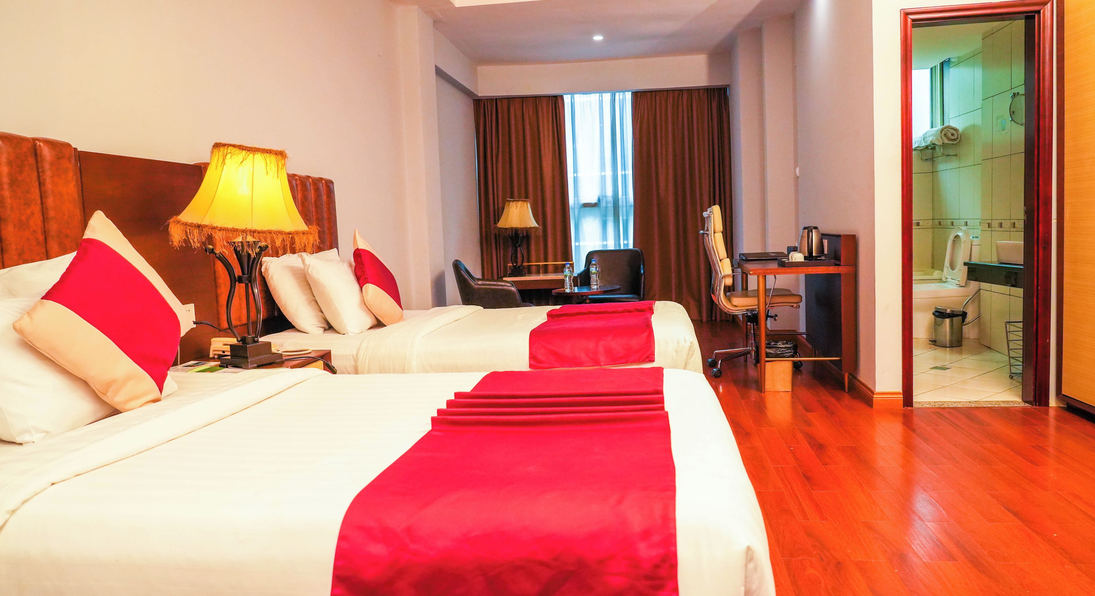 A hotel room with two beds, a desk, a chair, a lamp, and a bathroom visible through an open door.