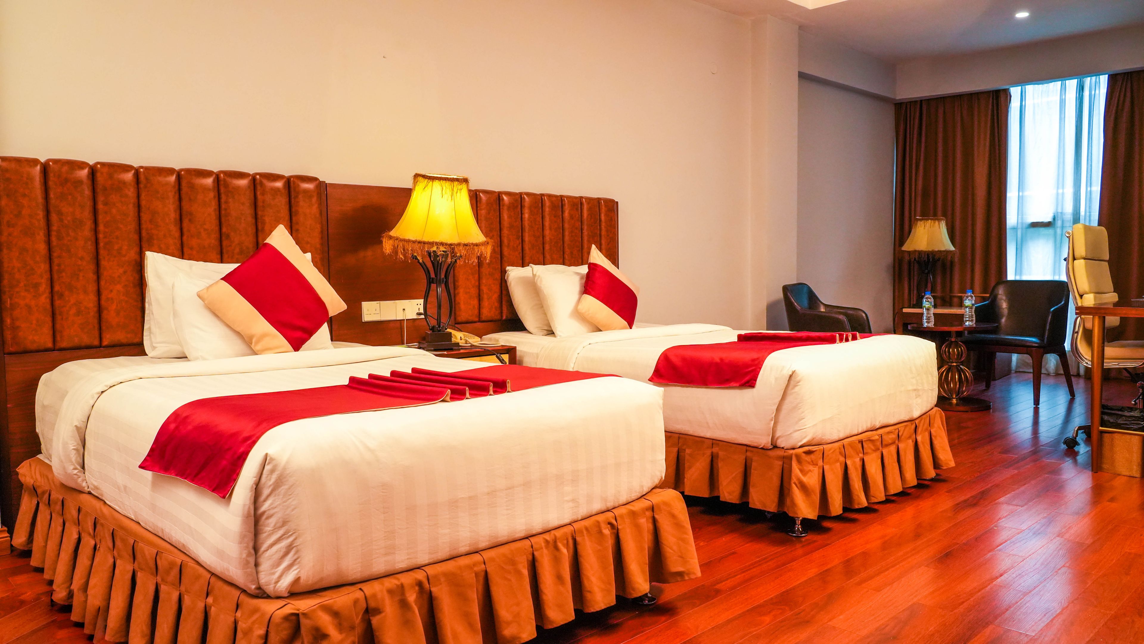 A hotel room with two beds, a wooden headboard, a desk with a chair, and a window with curtains.