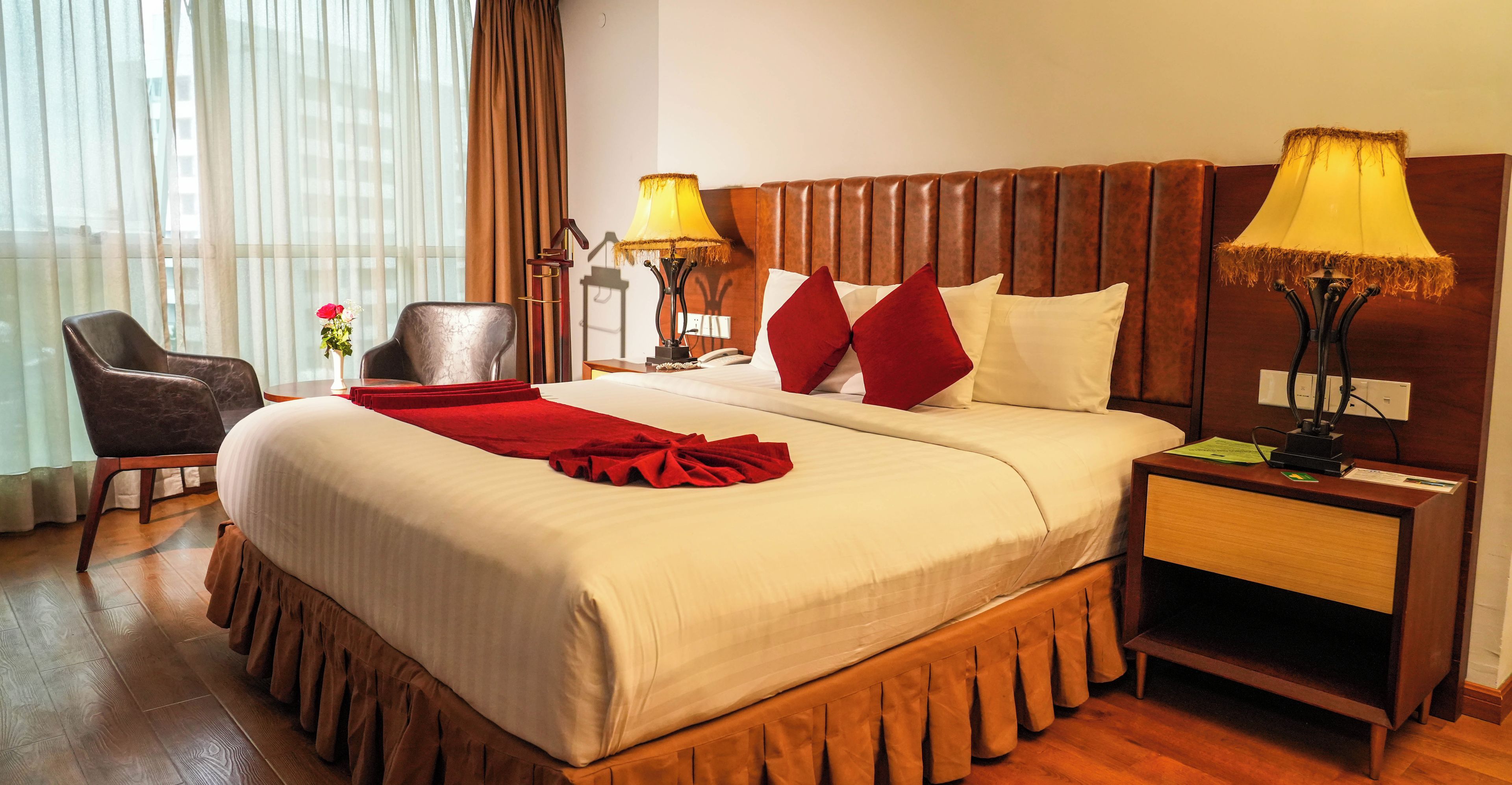 Une chambre d’hôtel avec un grand lit, deux lampes de chevet, une tête de lit en bois, une table de chevet, une chaise et une fenêtre avec des rideaux.