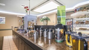 A hotel breakfast area with a granite countertop, coffee makers, a fruit basket, and a fresh sign.