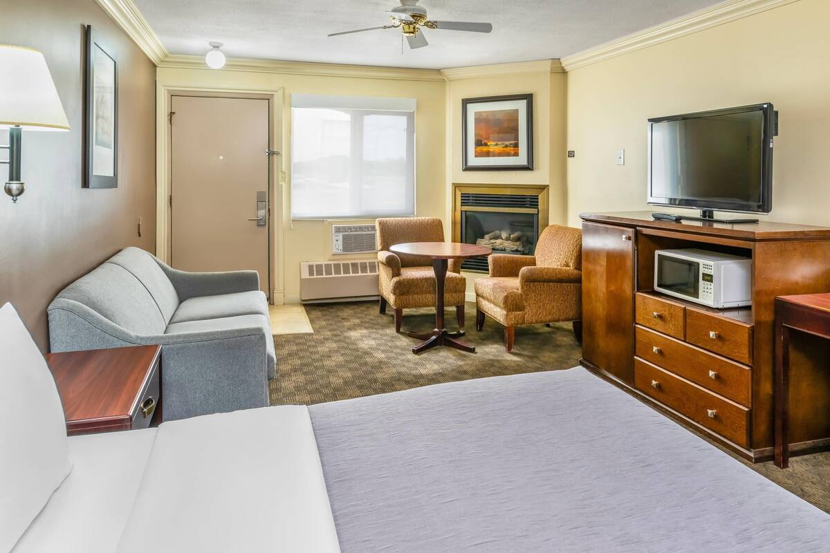 Quarto de hotel com cama, escrivaninha, televisão, lareira e área de estar com cadeiras.
