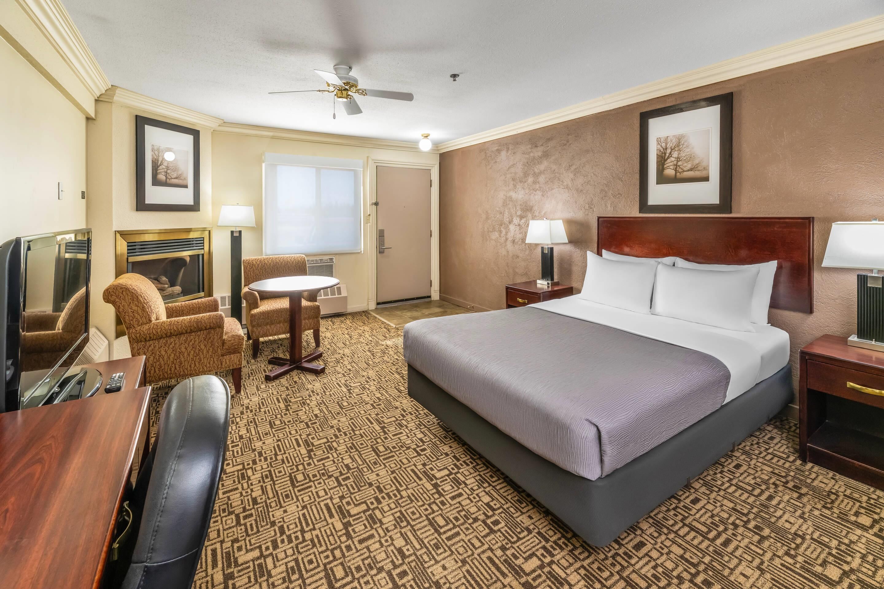 A hotel room with a large bed, a desk with a chair, a fireplace, a ceiling fan, and framed pictures on the walls.