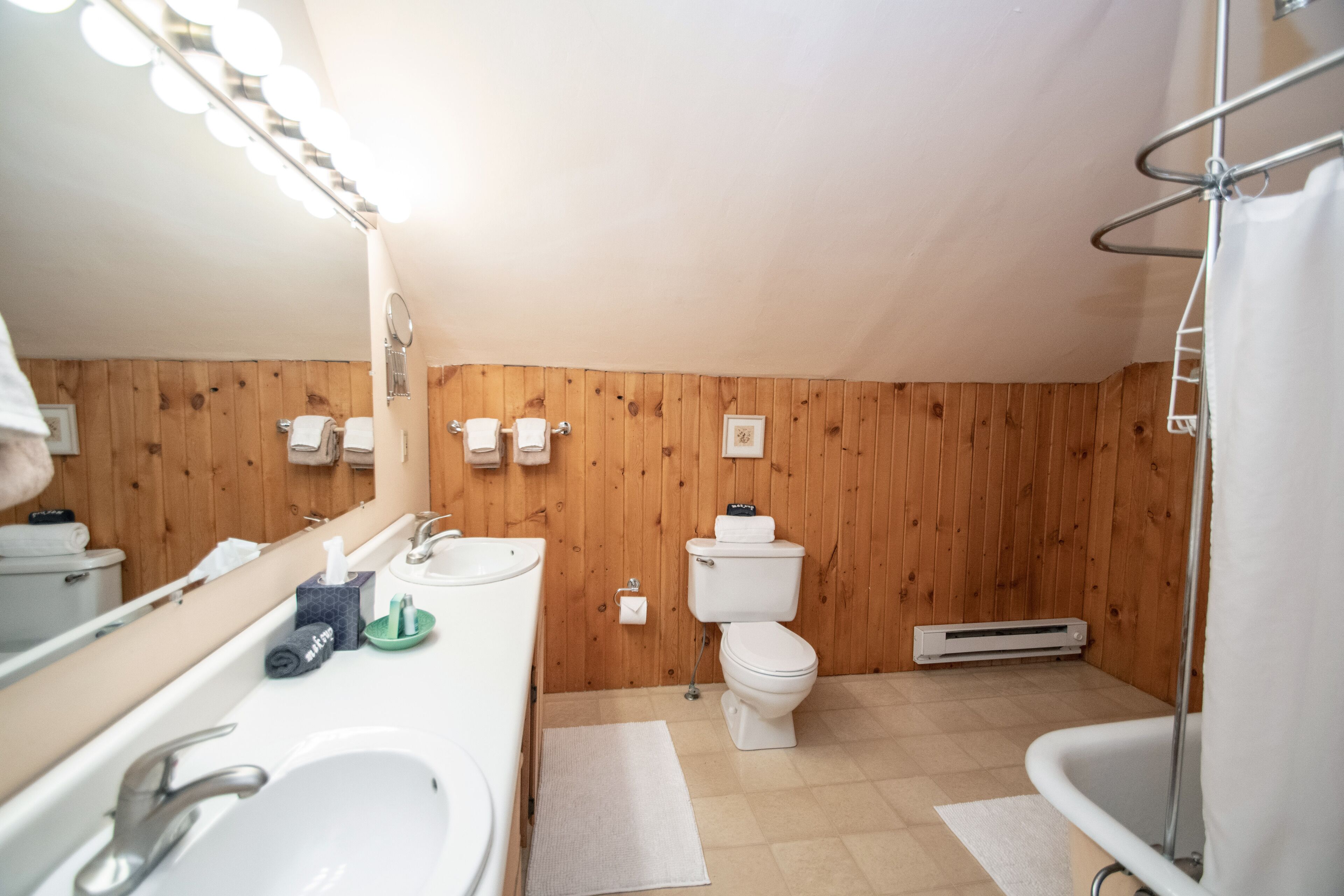 A bathroom with wooden paneling, a sink, a toilet, a mirror with lights, and a shower area.