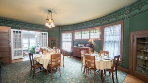 A dining room with a round table set for four, surrounded by wooden chairs. There is a chandelier hanging from the ceiling, and the room has patterned wallpaper and wooden paneling. - Looking Glass Inn (Indianapolis)