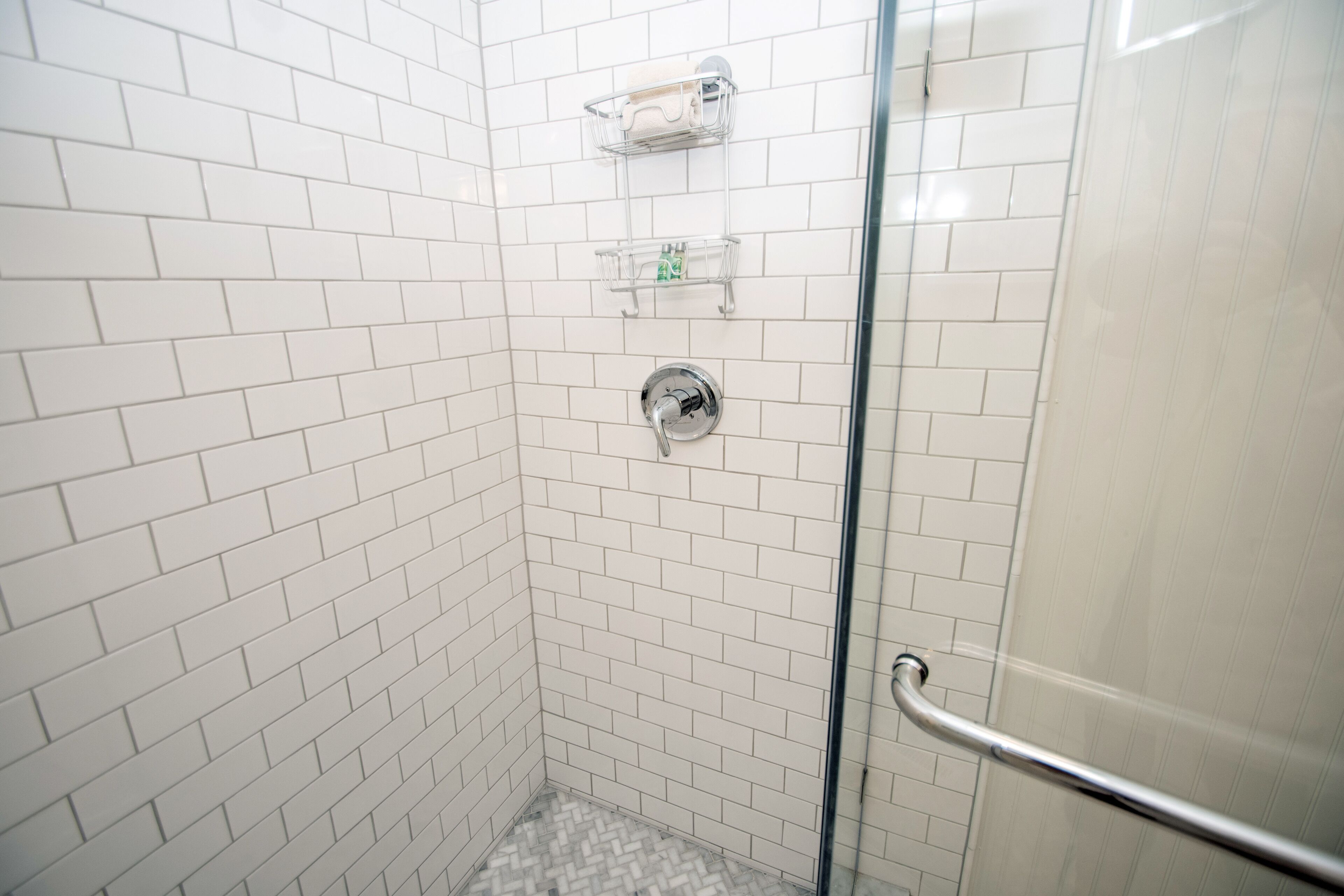 Un espace douche avec des murs recouverts de carreaux blancs, une porte en verre et une barre d’appui en métal.