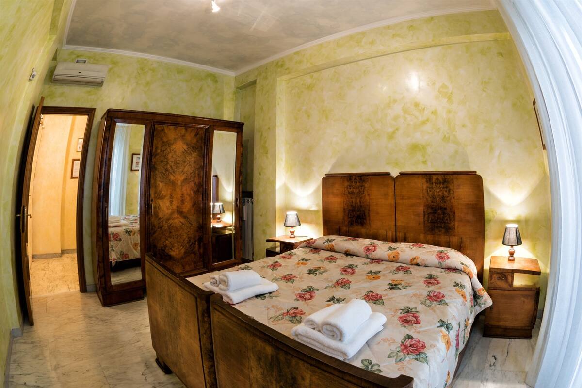 A bedroom with a wooden bed, two bedside tables with lamps, a mirror, and a door leading to another room.