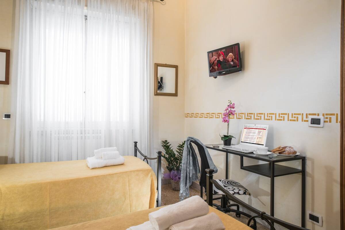 A hotel room with a bed, a desk with a laptop, a TV, and a window with curtains.