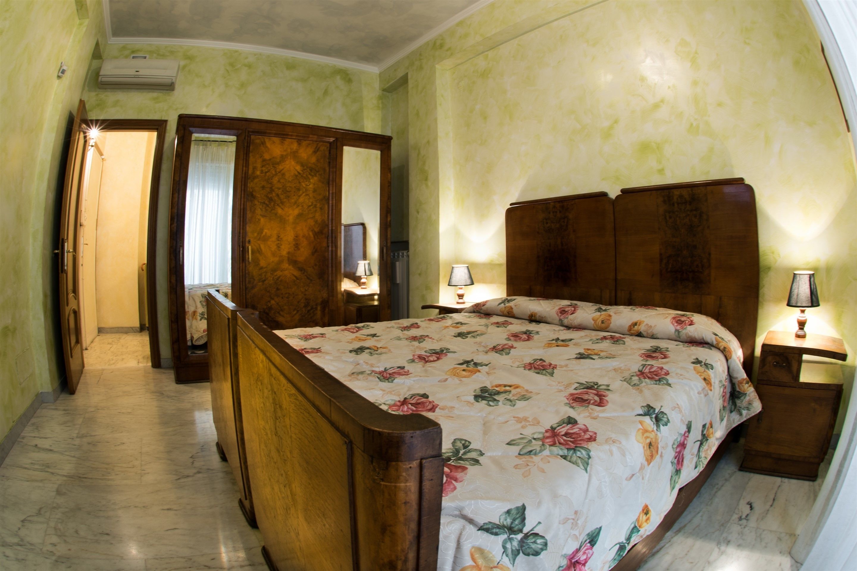 A bedroom with a wooden bed, floral bedding, a nightstand with a lamp, a mirror, and a door leading to another room.