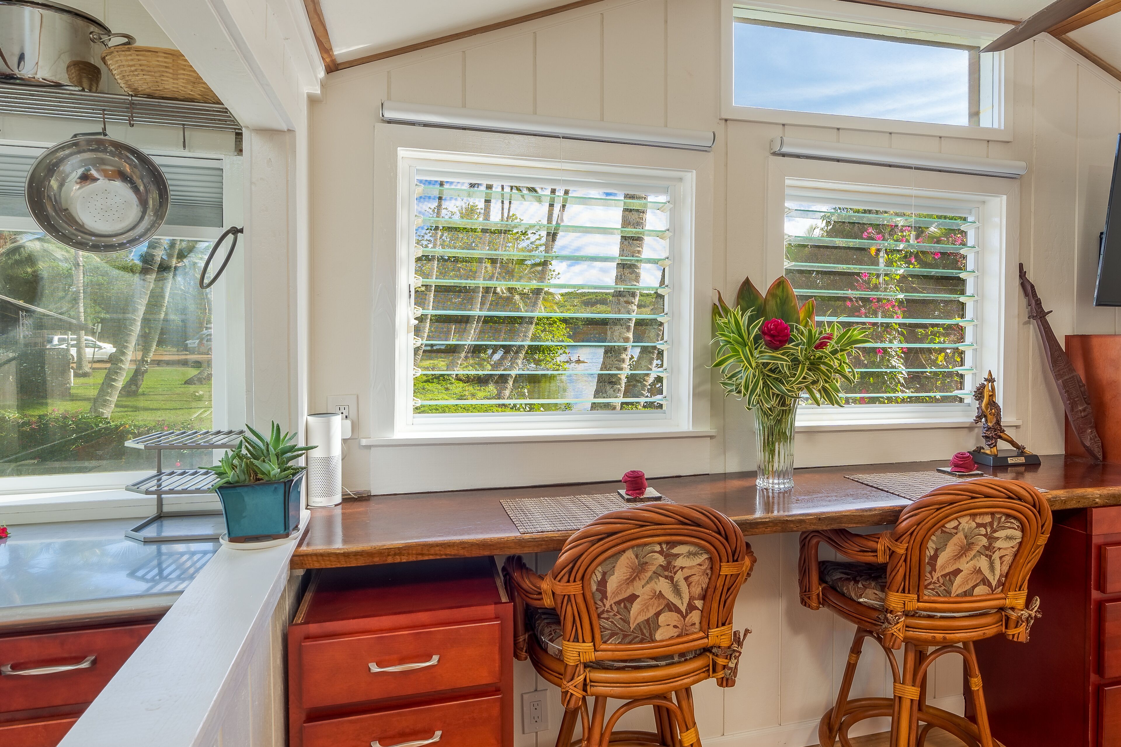 Una cocina con encimera de madera, dos sillas de mimbre, un florero con flores y vistas a la vegetación a través de las persianas.
