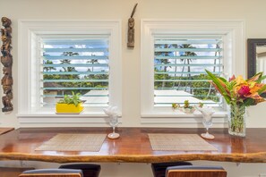 A dining area with a wooden table, two windows with blinds, a vase with flowers, and a small plant on the windowsill.