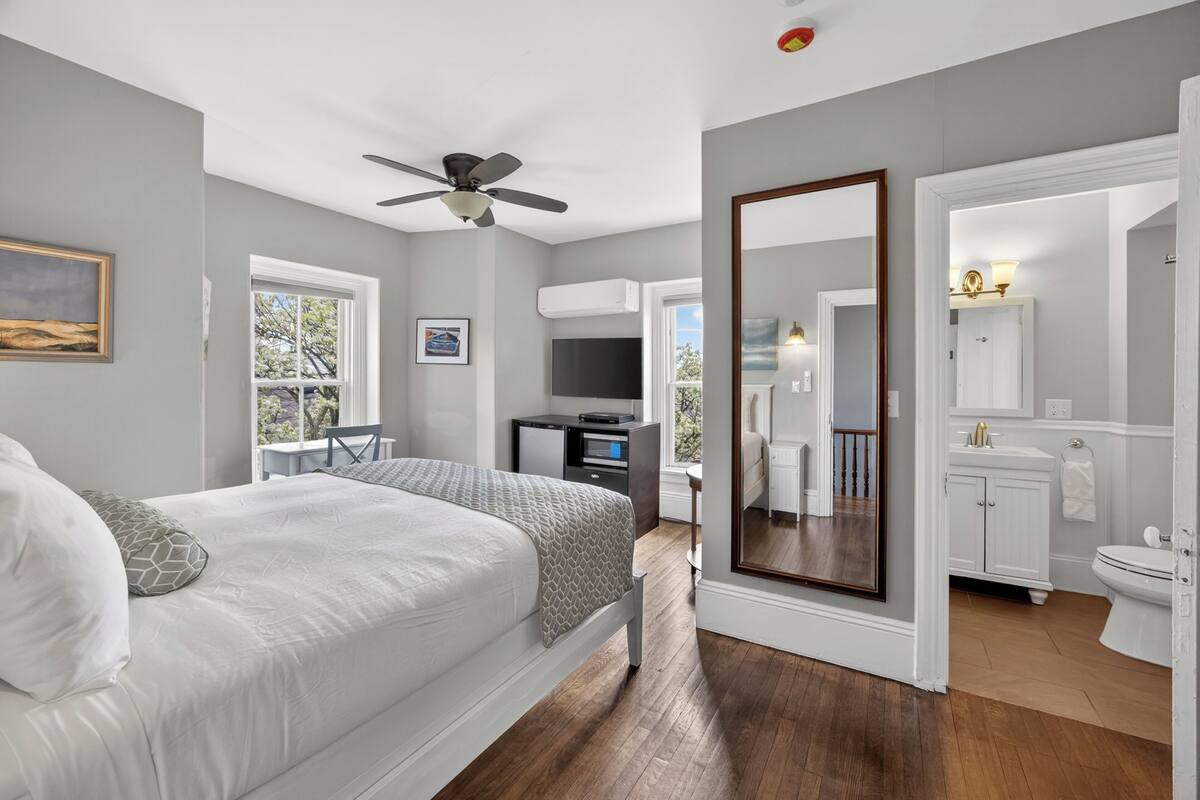 Une chambre à coucher comprenant un lit, un bureau, une chaise, un ventilateur de plafond, un grand miroir, ainsi qu’une salle de bain avec des toilettes et un lavabo.