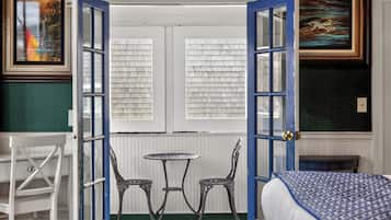 A room with a bed, a table with chairs, a window with shutters, and artwork on the walls.