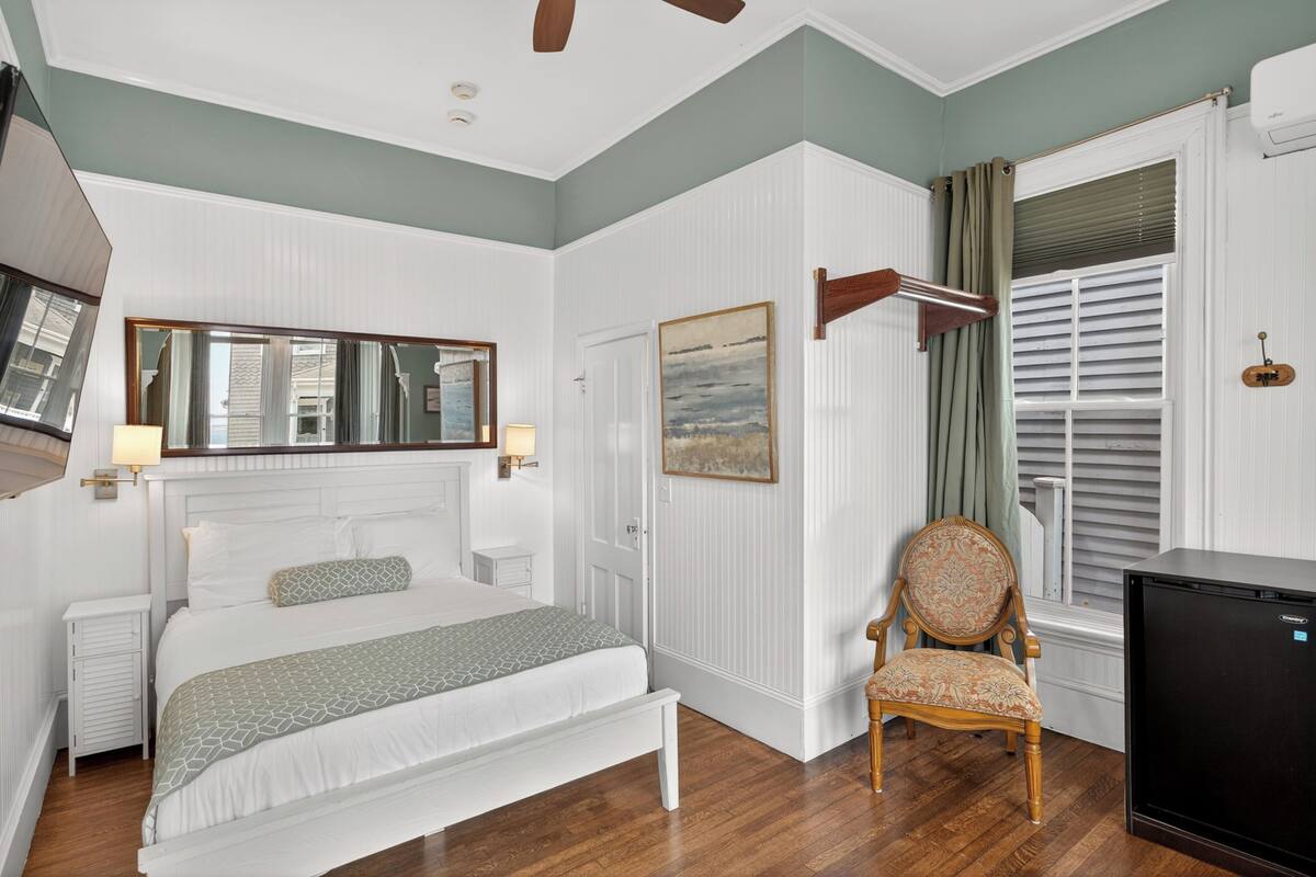 Une chambre à coucher avec un lit, une chaise, un bureau, un miroir et une fenêtre avec des stores.