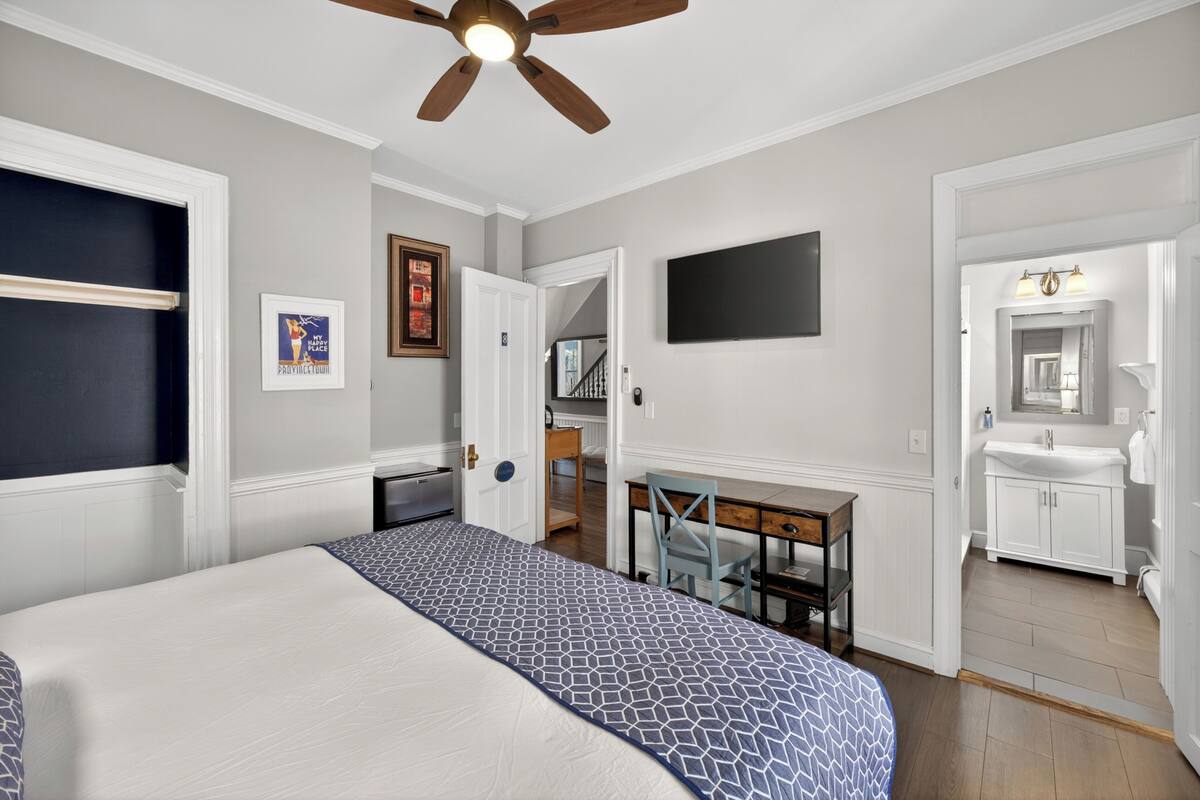 Une chambre à coucher comprenant un lit, un bureau, une chaise, un ventilateur de plafond, une télévision et une salle de bain avec un lavabo et un miroir.