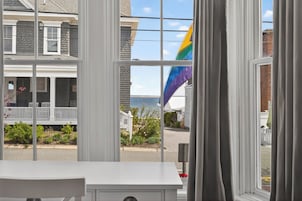 Vista a través de una ventana, con una mesa blanca en primer plano, una casa con porche y una bandera en el exterior.