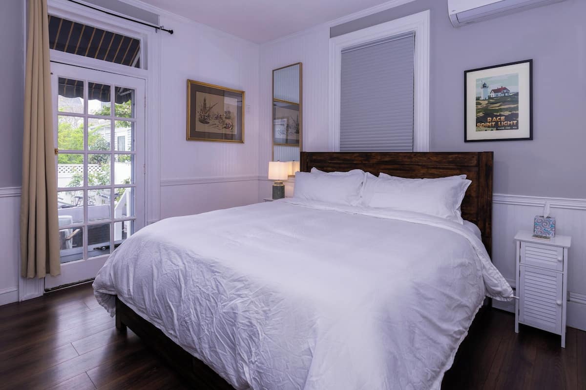 A bedroom with a large bed, a window with curtains, a bedside table, and a framed picture on the wall.