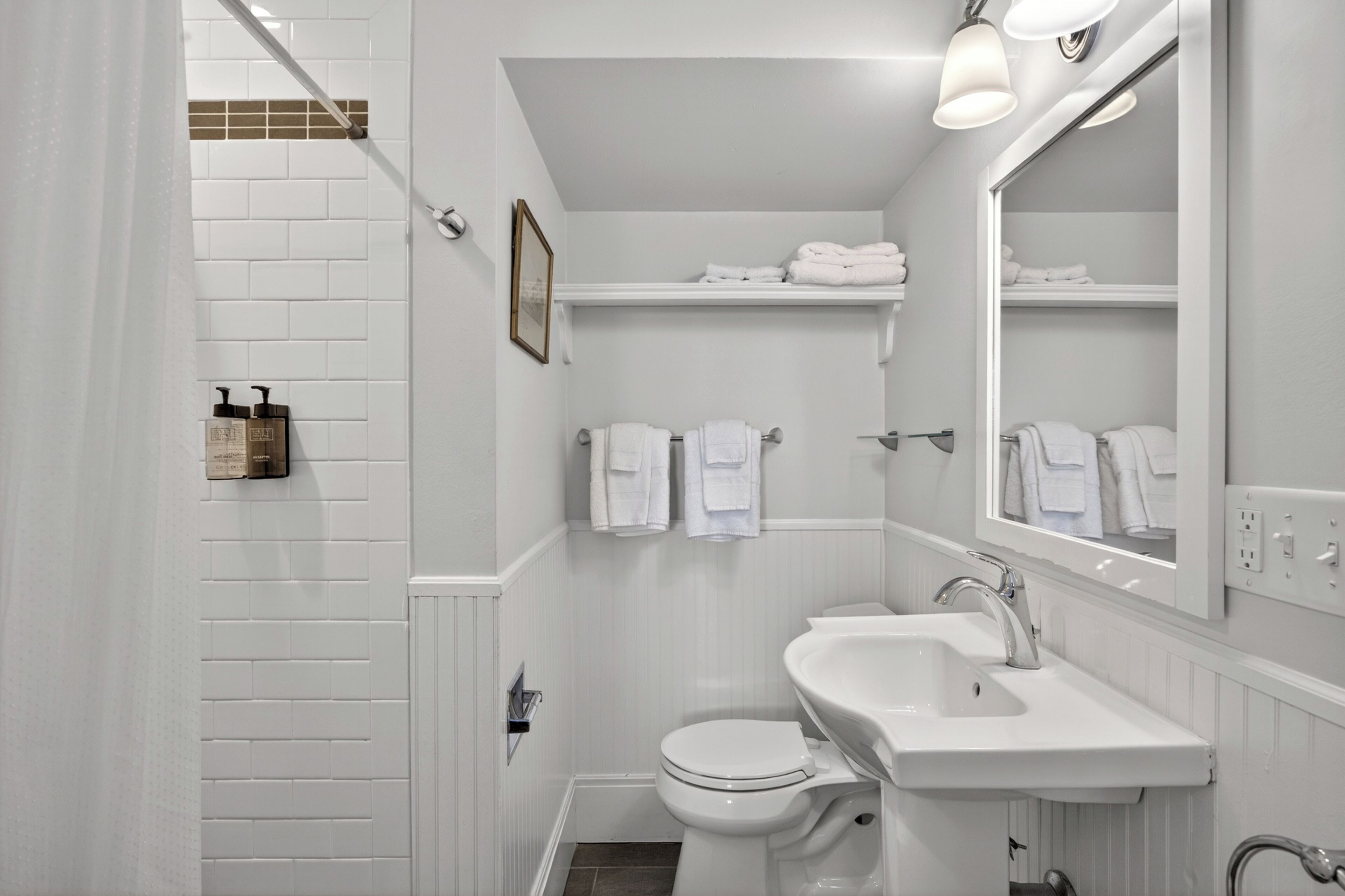 A bathroom with a white sink, toilet, and shower area. There are towels hanging on a rack and a framed picture on the wall.