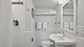 A bathroom with a white sink, toilet, and shower area. There are towels hanging on a rack and a framed picture on the wall.