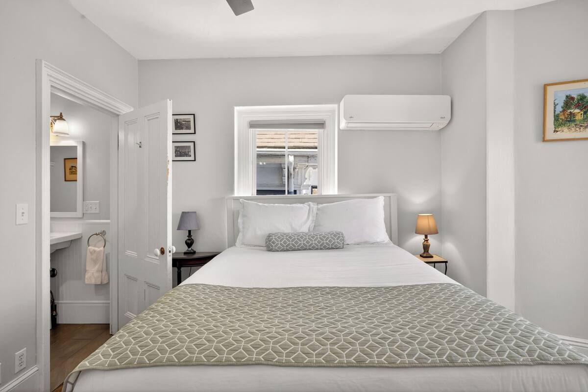Une chambre à coucher comprenant un lit, des lampes de chevet, une fenêtre, un appareil de climatisation et une porte donnant sur une salle de bain.
