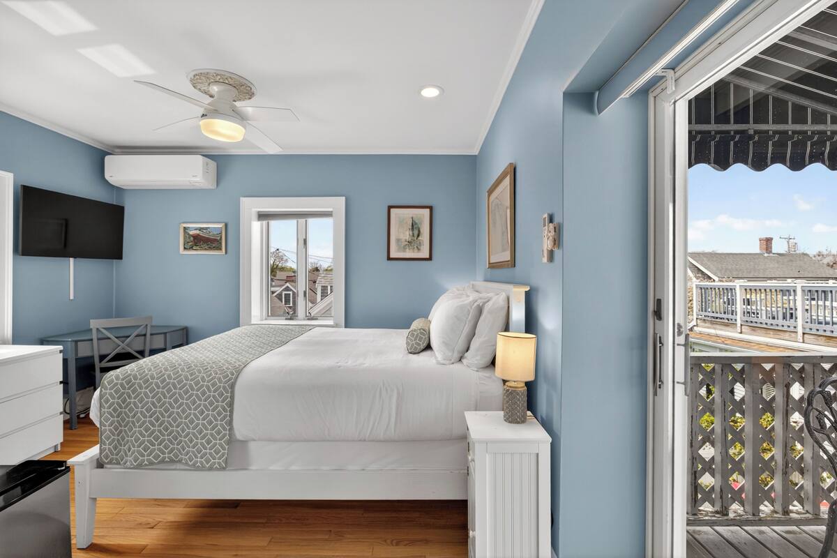 Une chambre à coucher comprenant un lit, une table de chevet, une lampe, un ventilateur de plafond, un appareil de climatisation et donnant sur un balcon.