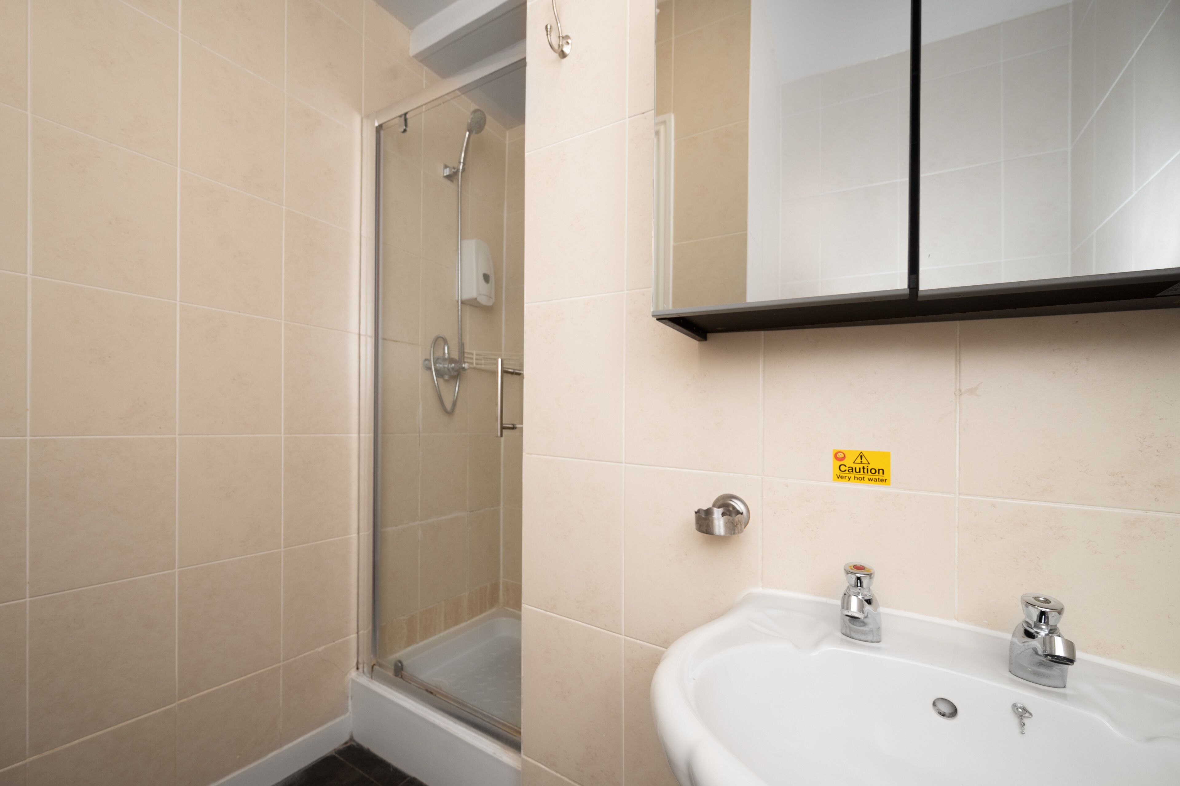 Un bany amb dutxa envidriada, un lavabo amb doble aixeta i un senyal de precaució a la paret.