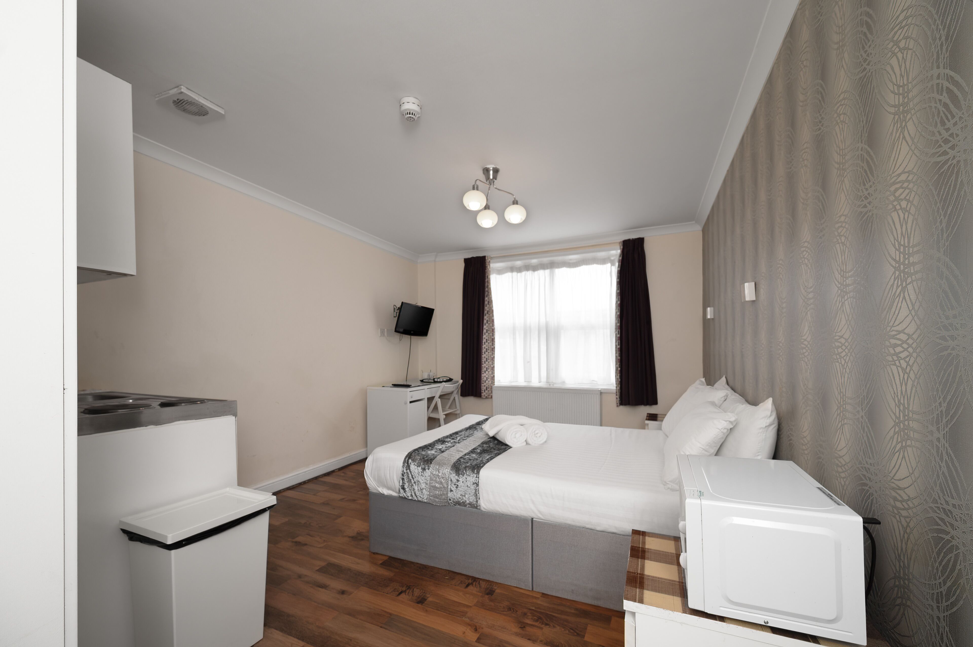 A bedroom with a bed, a desk, a chair, a microwave, and a small kitchenette.