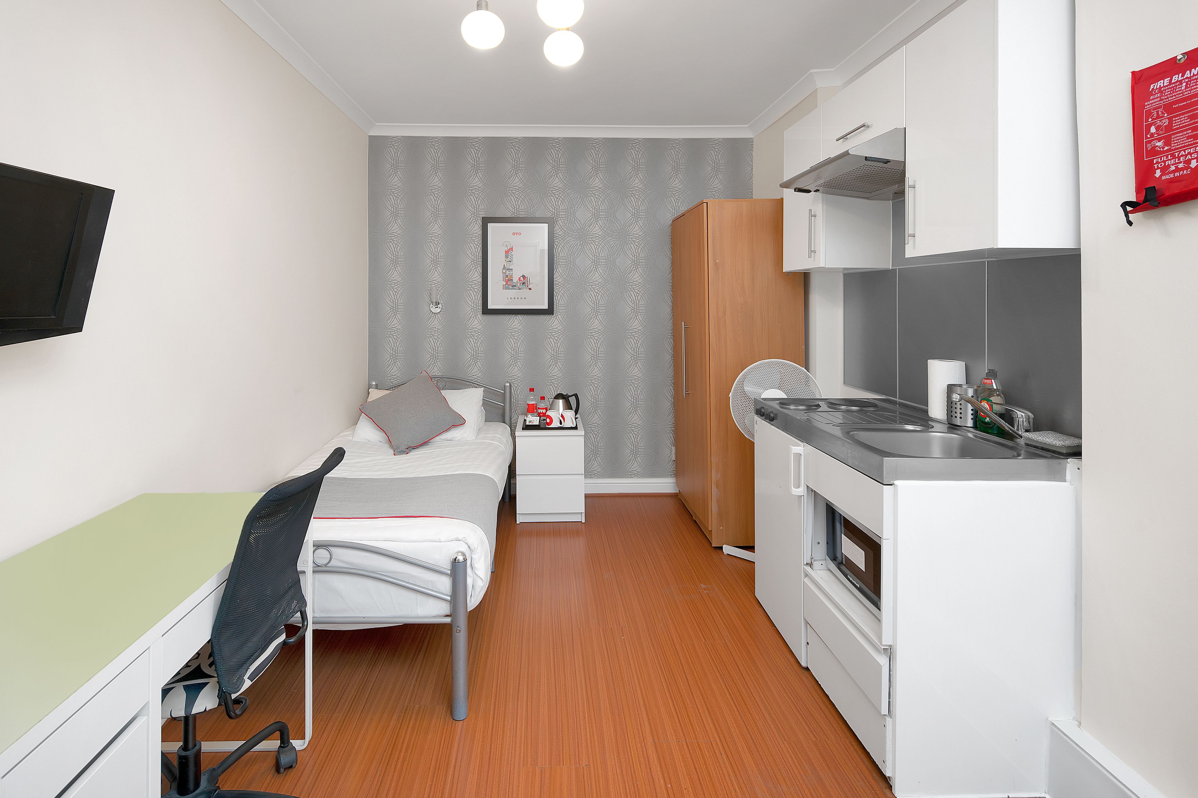 A compact hotel room with a single bed, bedside table, and a small kitchenette.