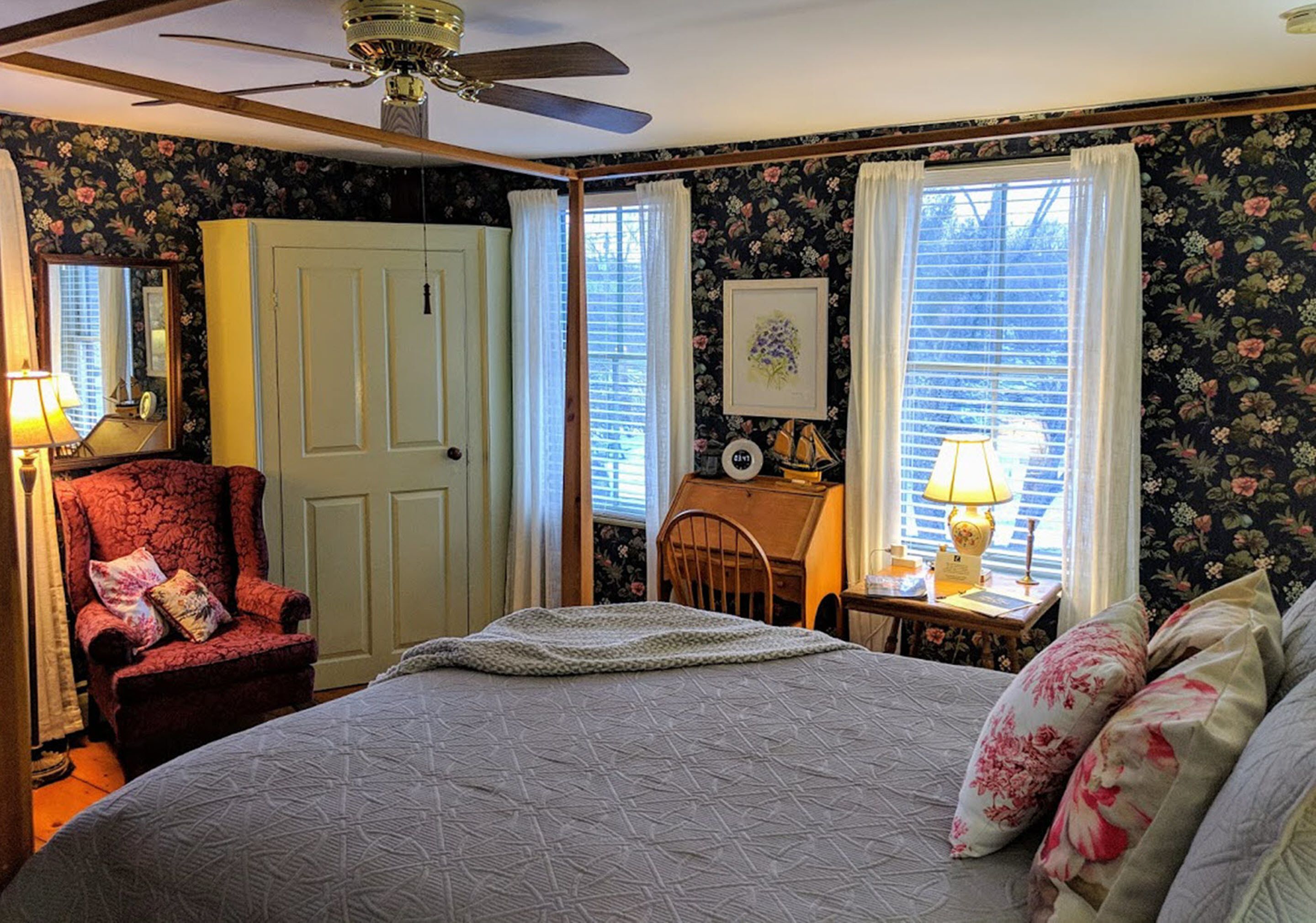 Quarto com cama, cadeira, escrivaninha, ventilador de teto e papel de parede floral.