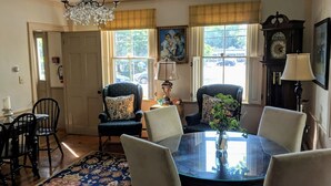 A dining room with a round table, four chairs, a chandelier, a clock, and a window with a view of the outside. - Elms of Camden (Camden)