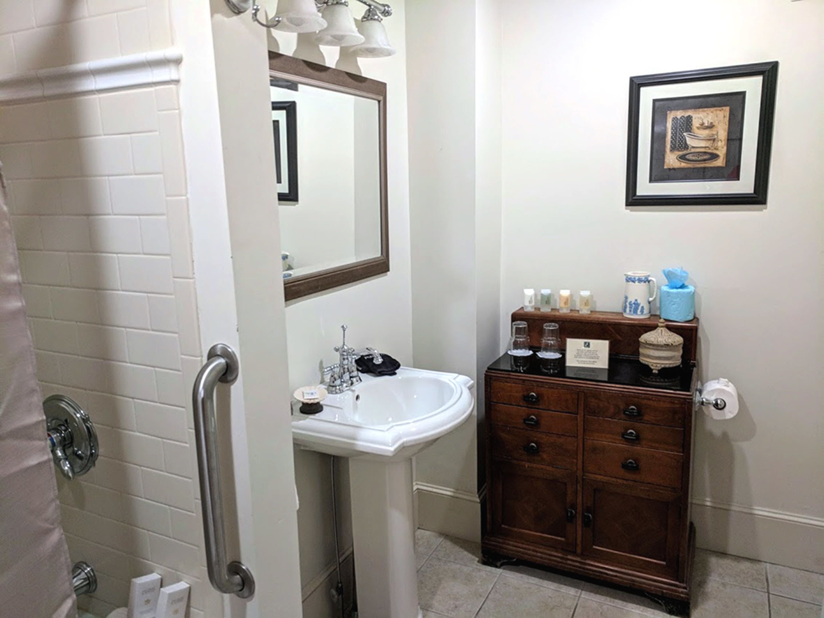 A bathroom with a white sink, a mirror, a wooden cabinet with toiletries, and a framed picture on the wall.