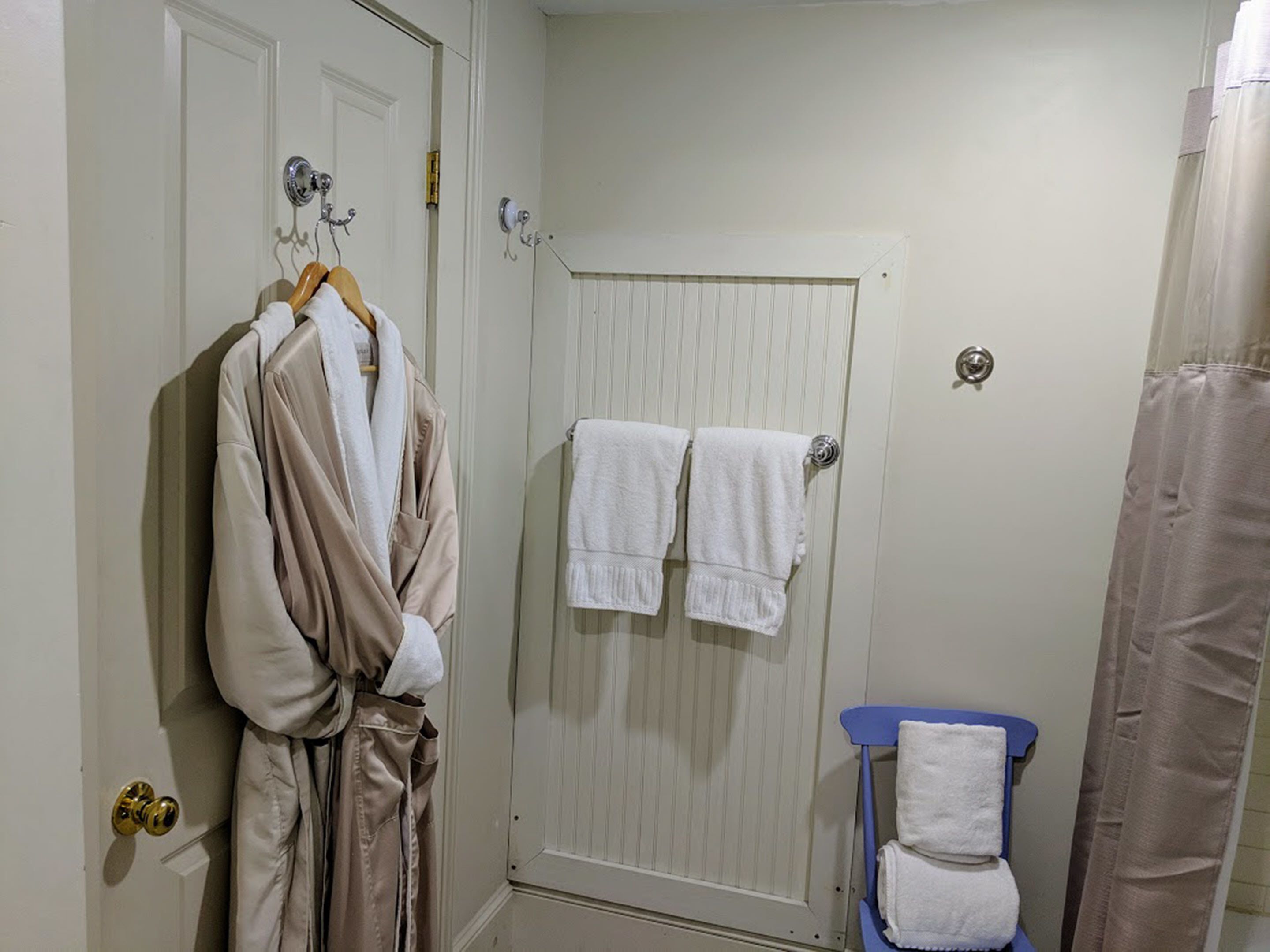 A bathroom with a white door, two towels hanging on a towel rack, a blue chair with a towel, and a shower curtain.