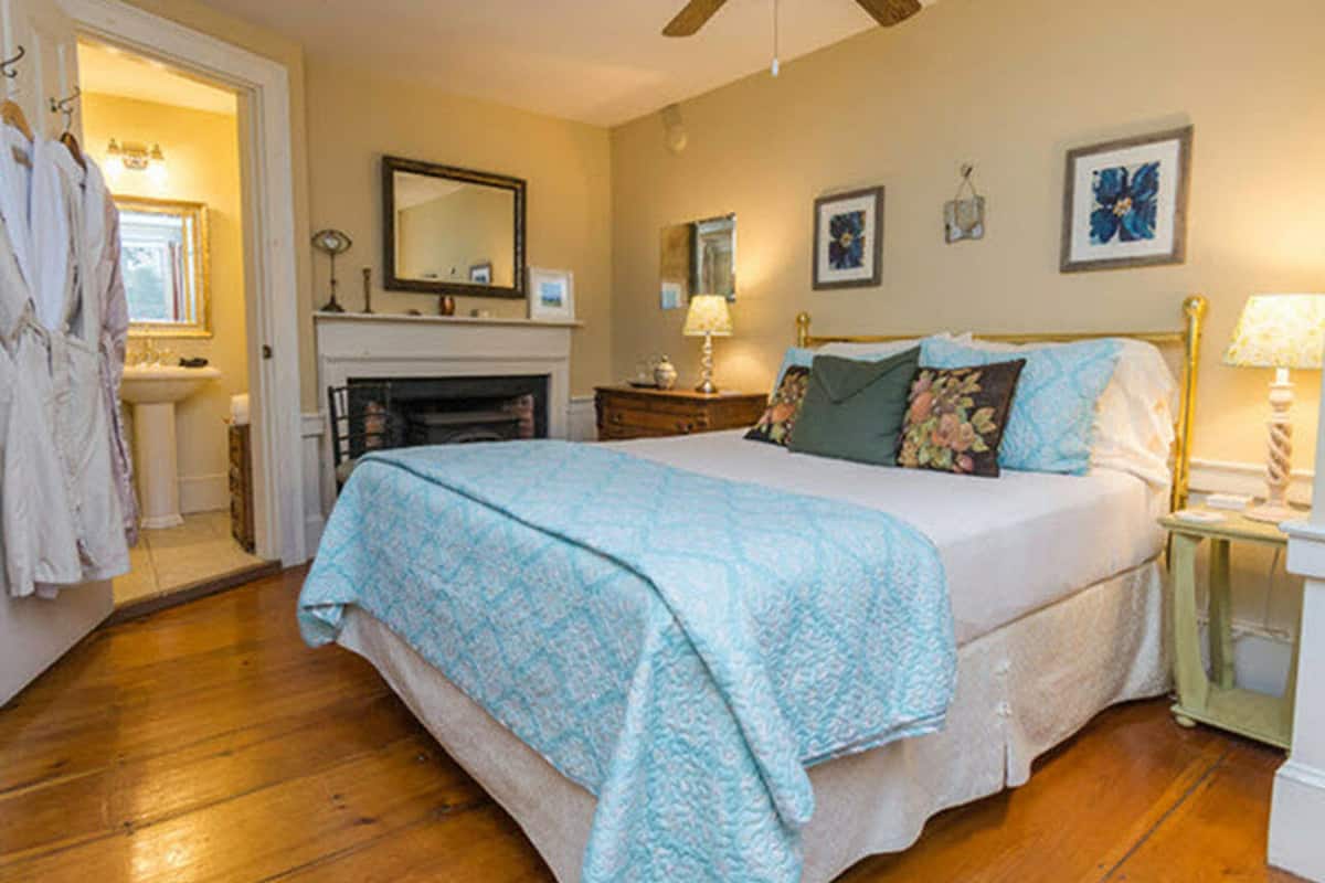 A bedroom with a bed, a nightstand, a dresser, a mirror, a fireplace, and a door leading to another room.