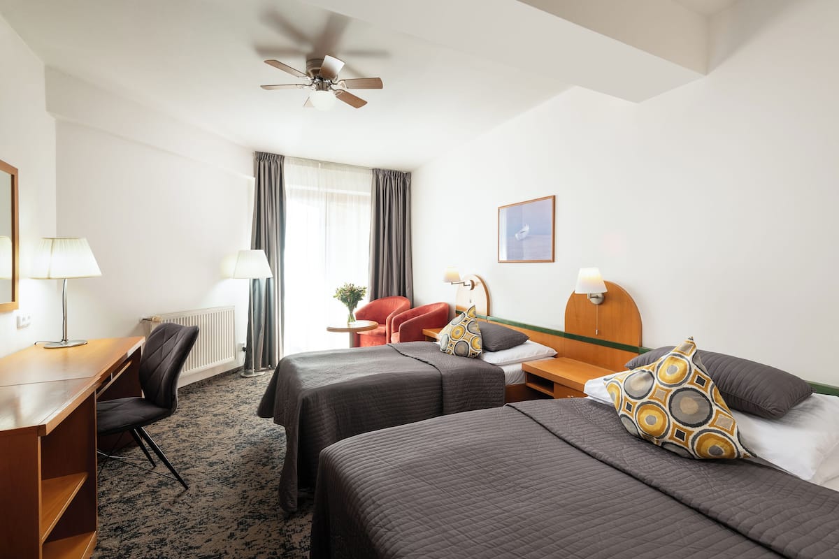 A hotel room with two beds, a desk, a chair, a ceiling fan, and a window with curtains.