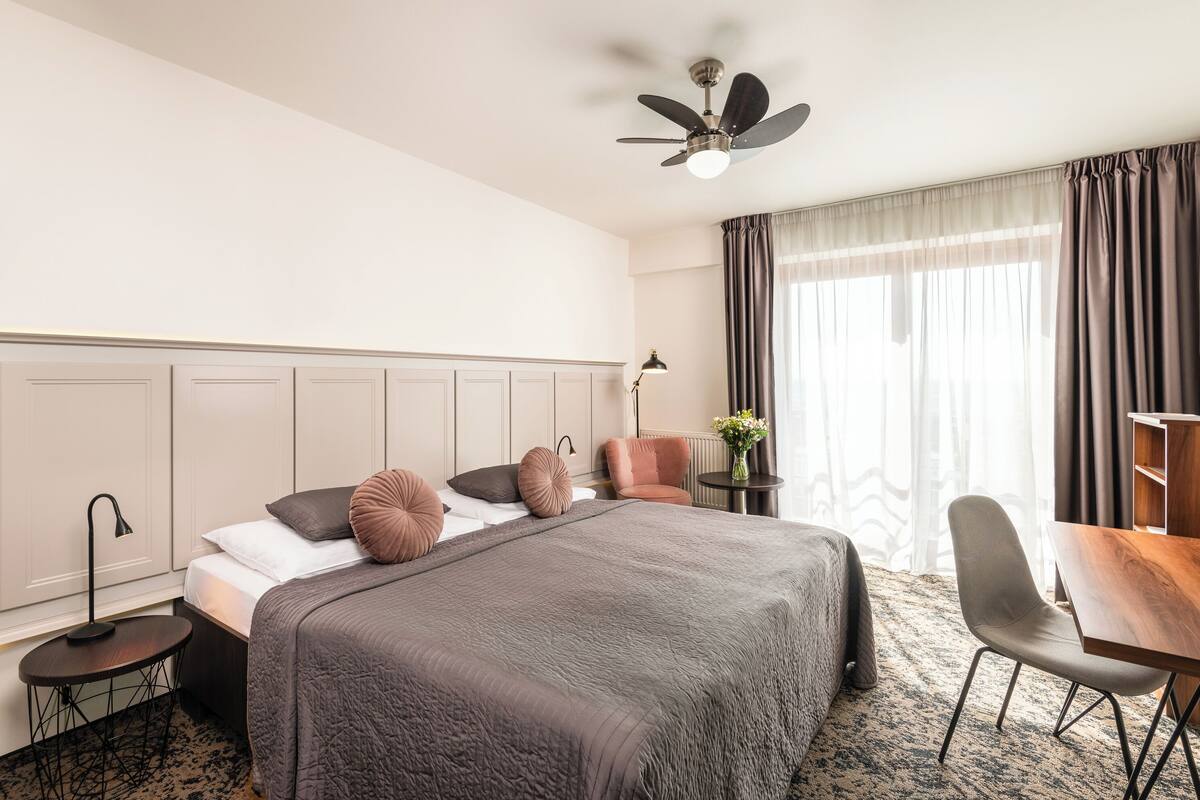 A hotel room with a large bed, a desk, a chair, a ceiling fan, and a window with curtains.