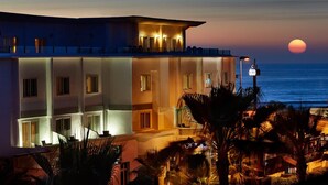 A hotel with a balcony overlooking the sea, featuring multiple windows and a well-lit facade.