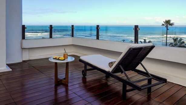 A balcony with a wooden floor, a lounge chair, and a small table with a drink and fruit.