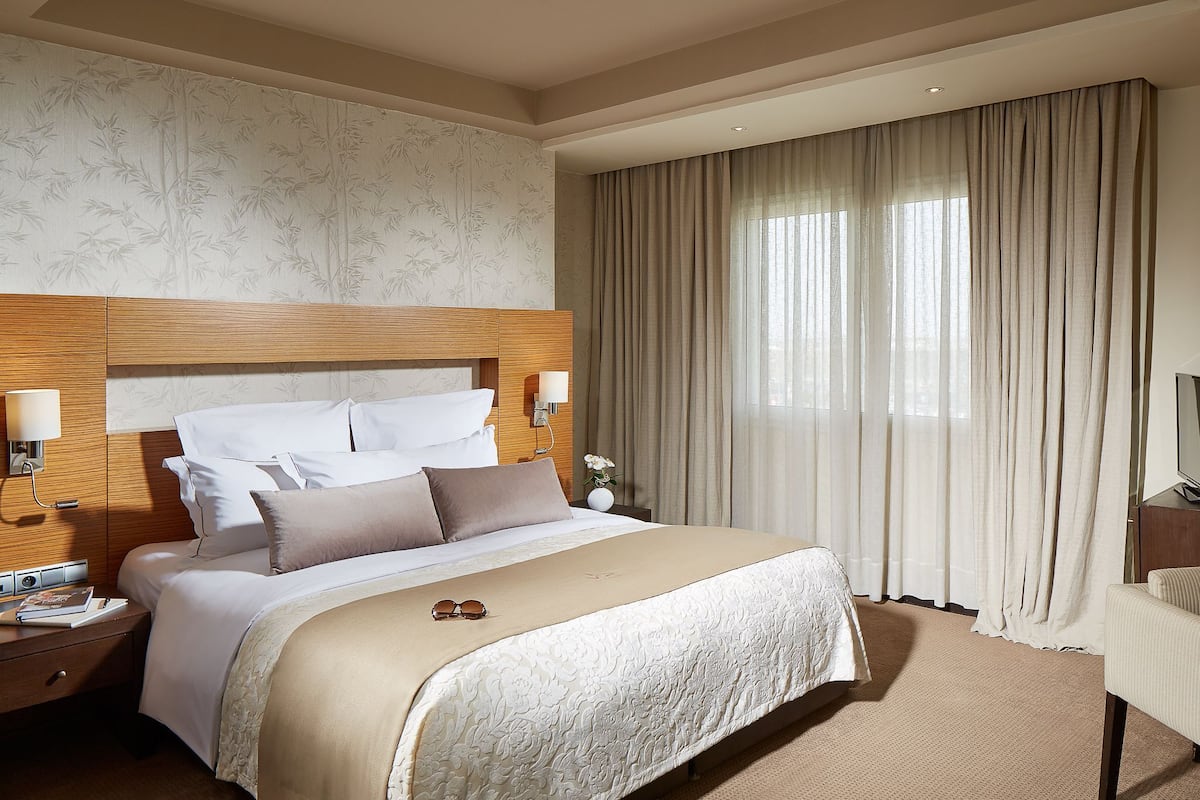 Una habitación de hotel con una cama grande, cabecera de madera, mesita de noche con lámpara y un ventanal con cortinas.