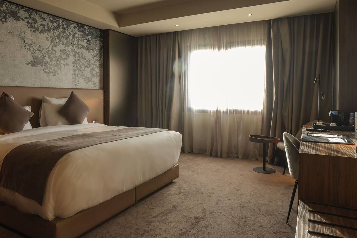 Habitación de hotel con una cama grande, un escritorio y vistas al exterior a través de cortinas translúcidas.