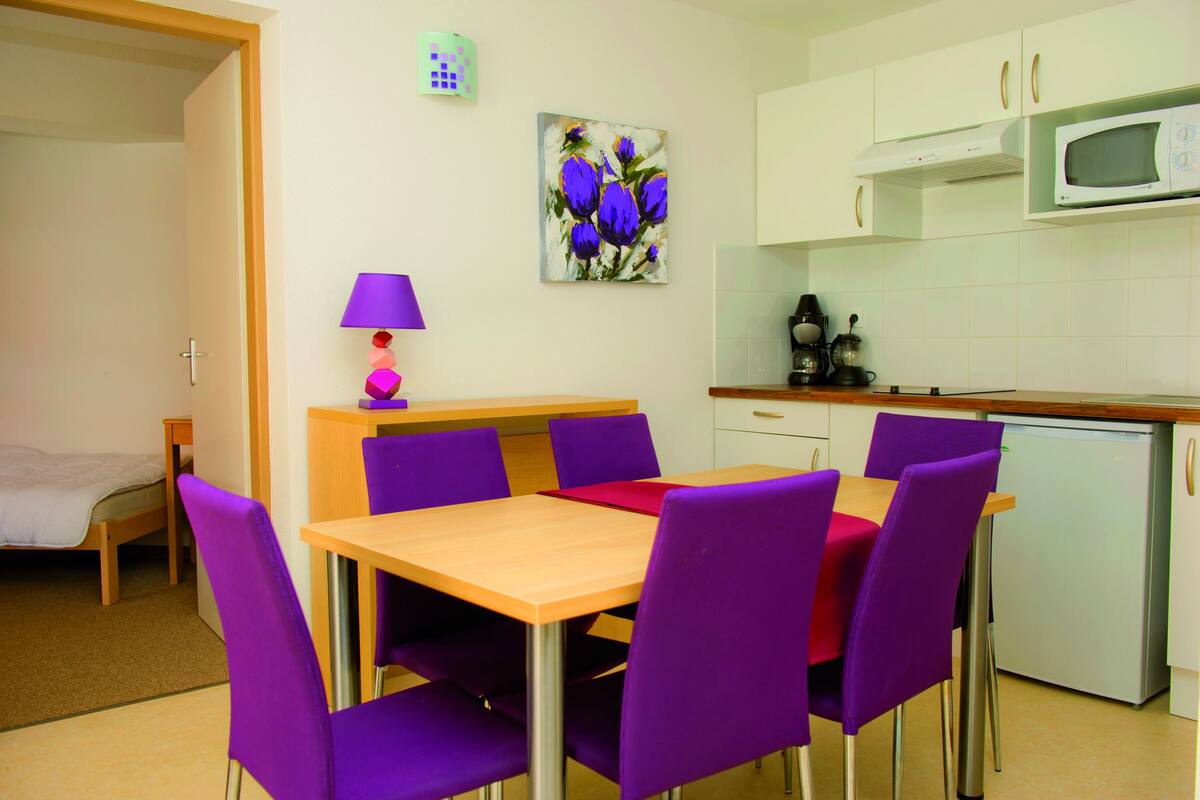 Cocina compacta con microondas, cafetera y refrigerador pequeño. Hay una mesa de comedor con sillas moradas y un cuadro de flores en la pared.