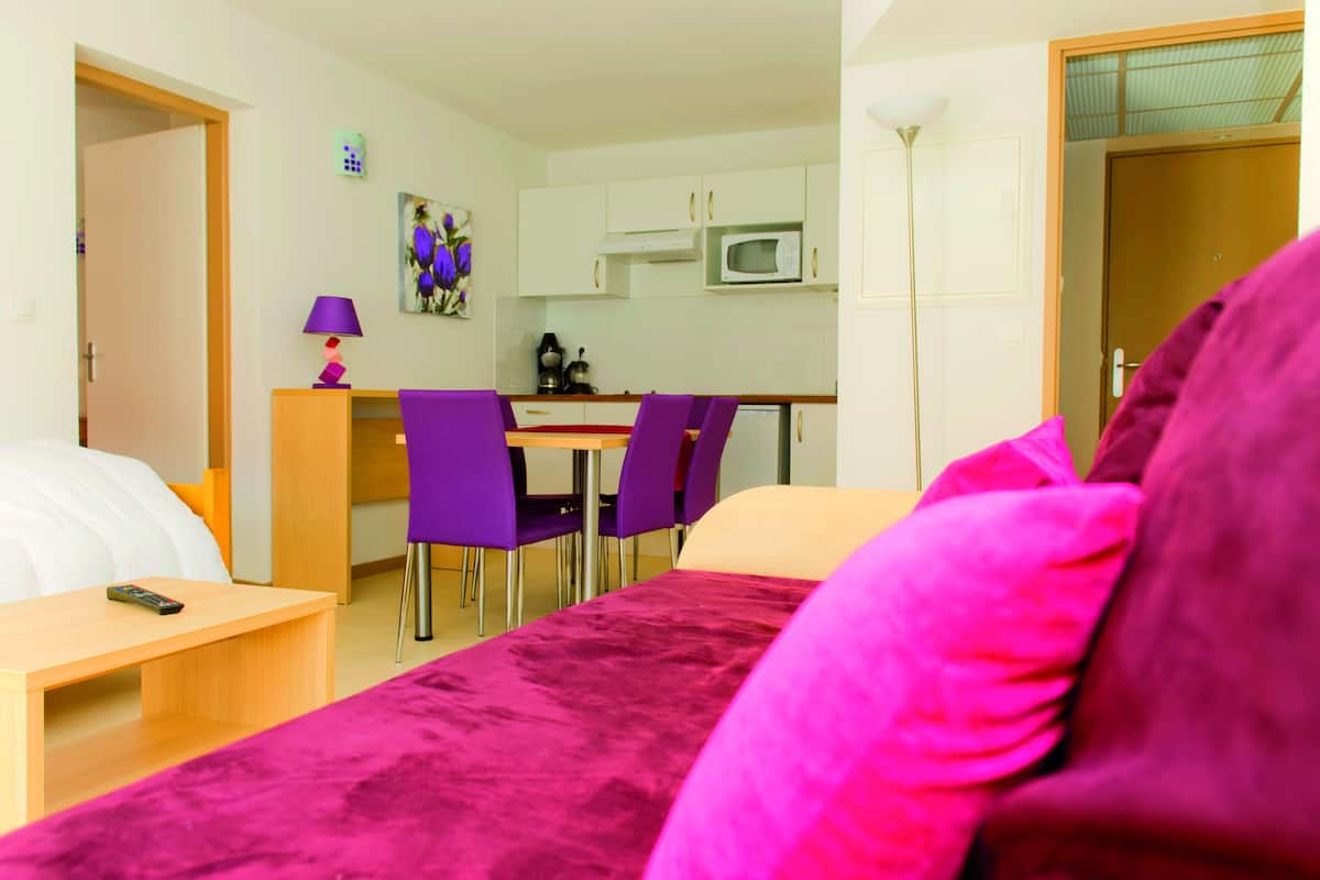 Una habitación de hotel pequeña con una cama, un escritorio, una silla, una mesa, un microondas, una lámpara y un cuadro en la pared.