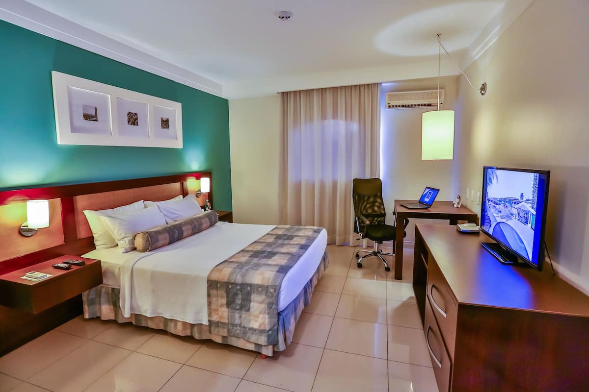 A hotel room with a bed, bedside table, lamp, TV, desk, chair, and curtains.