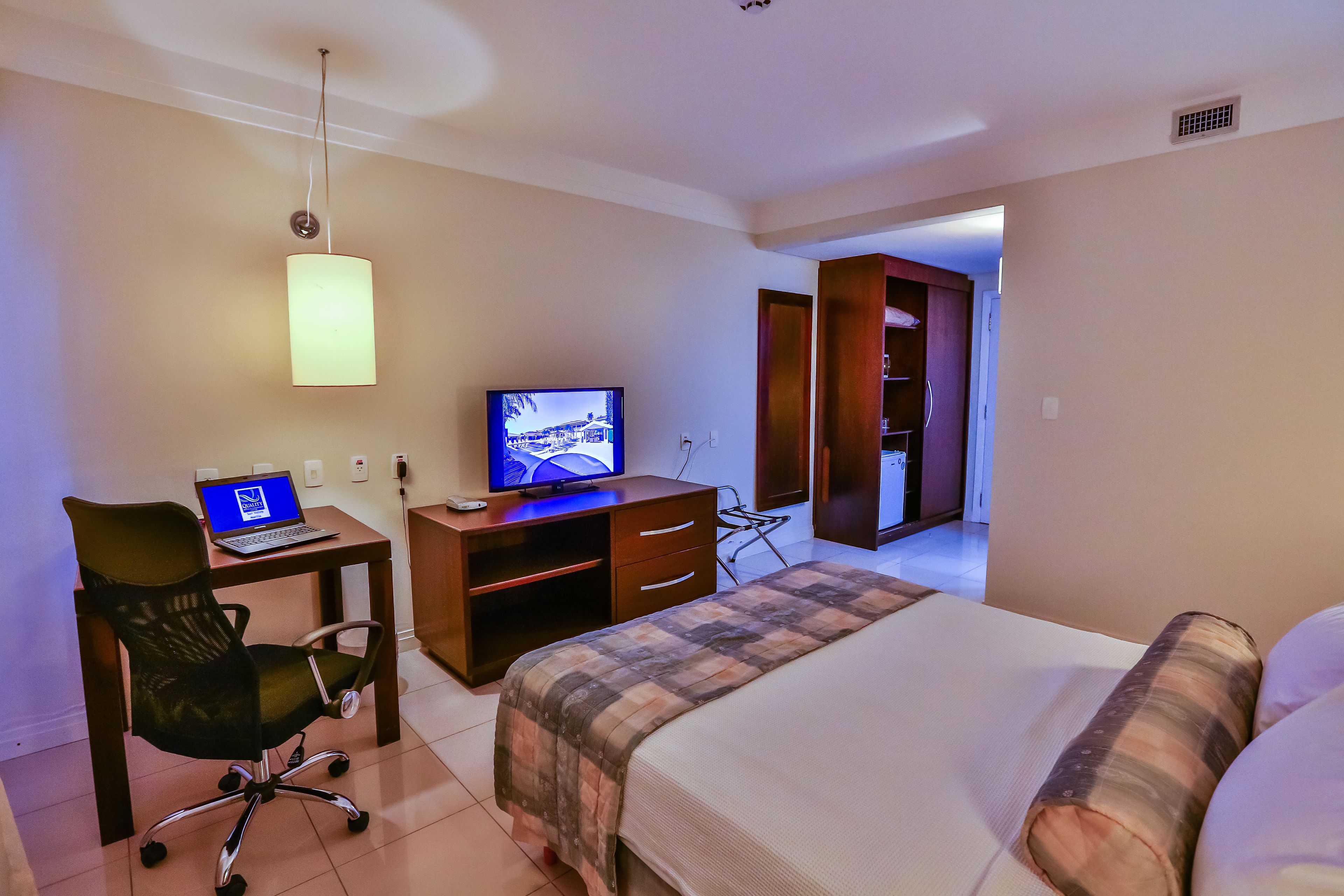 Una habitación de hotel con cama, escritorio, laptop, televisor, silla y armario.